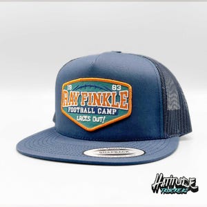 Pode incluir: Um chapéu de caminhoneiro azul e preto com um remendo que diz "Ray Finkle Football Camp Laces Out!" com os números 19 e 83 de cada lado do remendo.