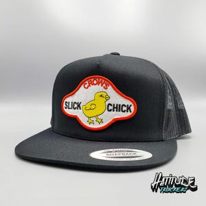 May include: Black trucker hat with a mesh back and a flat brim. The front features a patch with the text "CROW'S SLICK CHICK" around a yellow chick graphic. The hat has a snapback closure.
