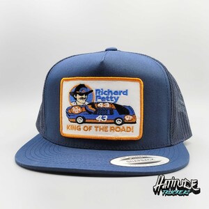 May include: A blue and black trucker hat with an embroidered patch featuring Richard Petty, the number 43, and the text "King of the Road!"