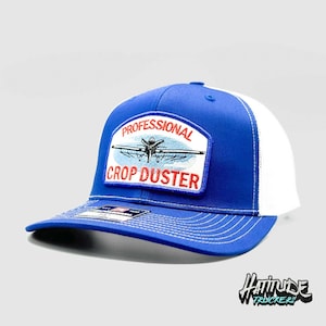 May include: A blue and white trucker hat with a patch that reads "PROFESSIONAL CROP DUSTER" with an image of a crop duster plane. The hat has a curved brim and a mesh back.