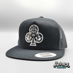 Puede incluir: Gorra de camionero negra con un símbolo de trébol negro y blanco y una malla negra en la parte posterior. La gorra tiene una etiqueta blanca "The Authentic Snapback" en la parte posterior.