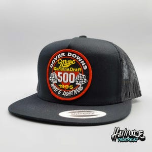 May include: Black trucker hat with a black mesh back. The front of the hat features a red, white, and yellow embroidered patch that says "Dover Downs Miller Genuine Draft 500 1995 White Lightning".