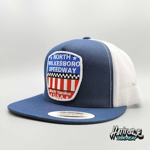 Può includere: Un cappello da camionista blu e bianco con una toppa che dice "North Wilkesboro Speedway" con un design di bandiera americana rosso, bianco e blu. La toppa dice anche "North Carolina" e "U.S.A."
