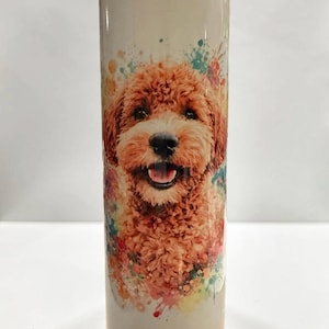 Peut inclure: Un gobelet cylindrique orné d'une illustration colorée style aquarelle représentant un chien au pelage bouclé brun doré. Le chien a une expression joyeuse, gueule ouverte, sur un fond de taches de peinture multicolores sur fond blanc.