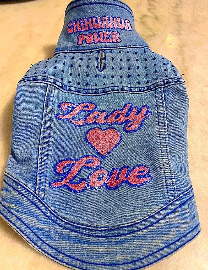 May include: A blue denim jacket with pink glitter lettering that says "Lady Love" and a pink glitter heart. The collar has blue glitter lettering that says "Chihuahua Power".