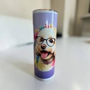 May include: A cylindrical stainless steel tumbler featuring a light purple background with a colourful watercolour splash design. The central graphic depicts a cream-coloured dog wearing black-rimmed glasses, with its tongue out in a happy expression.