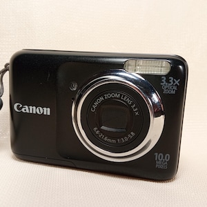 May include: A black Canon digital camera with a silver lens ring and a wrist strap. The camera has a 3.3x optical zoom and 10.0 megapixels. The Canon logo is visible on the front.