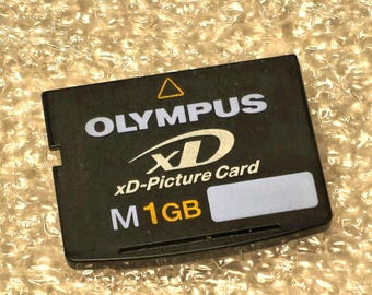 May include: A black Olympus xD-Picture Card memory storage device with white text reading OLYMPUS, xD, and xD-Picture Card. The card features a yellow triangle icon, a white M, and the text 1GB in yellow, along with a blank white label area.