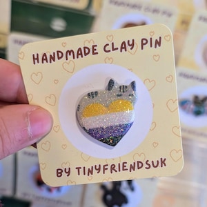 May include: A handmade clay pin shaped like a heart, featuring a glittery cat design. The pin incorporates the non-binary pride flag colours: yellow, white, purple, and grey. The card reads "HANDMADE CLAY PIN" and "BY TINYFRIENDSUK".