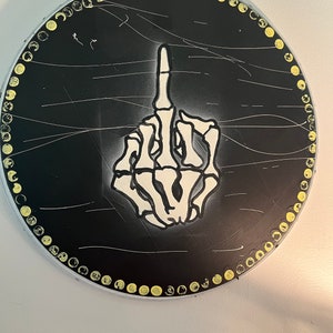 May include: A black circular canvas with a white skeleton hand giving the middle finger. The canvas is surrounded by a yellow circle of dots.