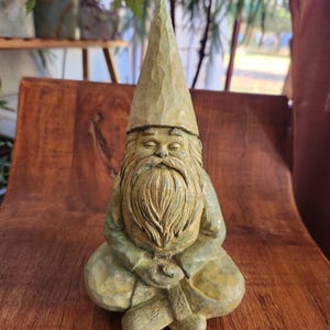 Könnte beinhalten: Eine Statue eines gnomes im Garten in Salbeigrün in einer sitzenden Meditationshaltung. Der Zwerg hat einen langen Bart, eine spitze Mütze und sitzt mit gekreuzten Beinen. Die Statue besteht aus einem strukturierten Material und ist ein dekoratives Gartenornament.