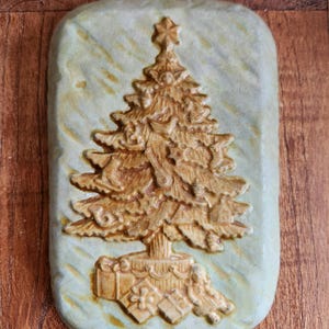 Puede incluir: Placa rectangular de cerámica con un relieve detallado de un árbol de Navidad. El árbol marrón tiene una estrella y regalos en la base, sobre un fondo verde pálido texturizado. Un artículo decorativo festivo para la temporada navideña.