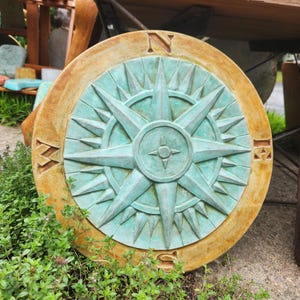 May include: A circular, weathered compass rose in teal and brown tones. The compass has directional markers, including "N" and "S", and a central starburst design. The outdoor decor piece is surrounded by green foliage.