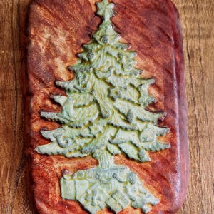 Puede incluir: Adorno navideño rectangular de estilo rústico. Presenta un diseño detallado de árbol de Navidad verde claro con una estrella. El árbol se encuentra sobre un fondo texturizado marrón rojizo, creando una decoración festiva.