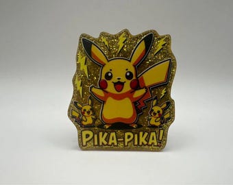 Puede incluir: Una placa decorativa dorada y brillante que presenta un personaje Pikachu amarillo grande en el centro, flanqueado por dos figuras Pikachu más pequeñas. El diseño incluye rayos amarillos y el texto PIKA-PIKA! en letras amarillas.