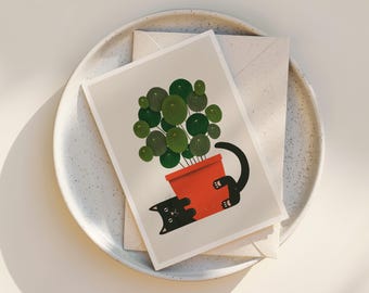 May include: A greeting card featuring a whimsical illustration of a black cat playfully interacting with a potted plant. The plant has large, round green leaves and is in a bright red pot. The card rests on a speckled white plate with an envelope.