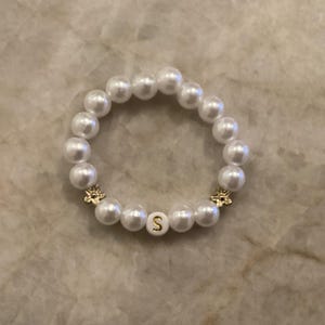 May include: White pearl bracelet with gold accents. The bracelet features large, round, white pearls strung together. A gold-colored charm and a bead with the letter "S" add detail to the design. The bracelet is a stylish accessory.