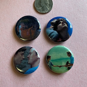 以下が含まれることがあります： 映画「君の名前で僕を呼んで」の画像が描かれた丸いボタンが4つ。最初のボタンは、エリオのアップ。2つ目は、エリオとオリバーが空を見上げている。3つ目は、エリオとオリバーが互いに見つめ合っている。4つ目は、エリオとオリバーがビーチを歩いている。