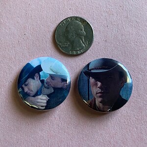 May include: Two round button pins featuring a black and white photo of two men wearing cowboy hats. The men are looking at the camera and appear to be in a close embrace.