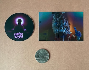 May include: Two stickers, one round and one rectangular. The round sticker has a purple glow and the text "I saw the glow" in white. The rectangular sticker shows a creature with a blue and green iridescent body and a blue hand holding a blue object.