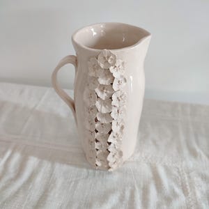 Peut inclure: Une cruche en céramique rose pâle avec une anse et un bec verseur. La cruche est décorée d'une rangée verticale de motifs floraux blancs en relief. La cruche est posée sur une surface blanche.