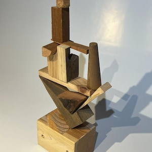 May include: A vertical sculpture constructed from various stacked wooden blocks and shapes in different shades of brown and tan. The structure features a rectangular base, a triangular middle section, and several smaller geometric pieces on top.