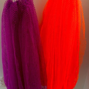 May include: Two mesh bath poufs, one vibrant purple and the other bright orange, are suspended by their drawstring loops. The poufs are made of a loosely woven material, and are hanging against a white wall.