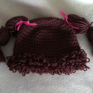 May include: A brown crocheted baby hat designed to resemble a doll's hairstyle. The hat features two yarn pigtails tied with pink ribbons and a fringe of curly yarn around the brim. This handmade hat is a unique accessory for infants.