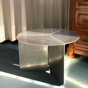 May include: A modern, brushed metal side table with a circular top and three supporting panels. The table is approximately 24 inches in diameter and 18 inches tall. The design is minimalist and industrial, suitable for contemporary interiors.