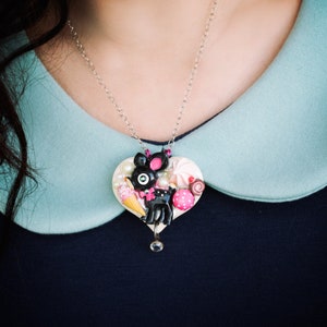 Puede incluir: Un collar con un colgante en forma de corazón blanco y negro con un amuleto de conejo negro, caramelos rosas y blancos y una perla. El collar está en una cadena plateada.