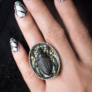 May include: A silver ring with a black beetle design surrounded by green and gold rhinestones. The ring is on a hand with black and white nail polish.