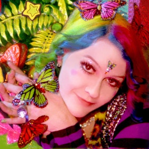 May include: A woman with rainbow hair and a butterfly sticker on her forehead. She is wearing a purple and black striped shirt and has a butterfly on her finger.