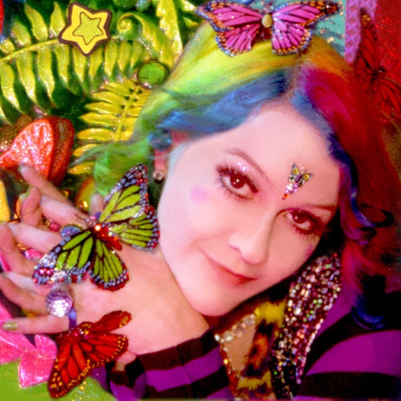 May include: A woman with rainbow hair and a butterfly sticker on her forehead. She is wearing a purple and black striped shirt and has a butterfly on her finger.