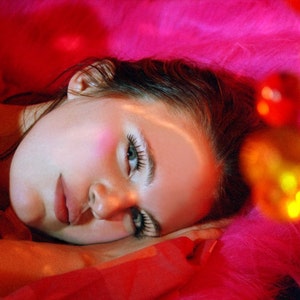 May include: A woman with dark hair lies on a pink fuzzy surface. Her eyes are closed and she is wearing makeup. The background is out of focus and features a string of colorful lights.