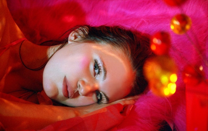 May include: A woman with dark hair lies on a pink fuzzy surface. Her eyes are closed and she is wearing makeup. The background is out of focus and features a string of colorful lights.