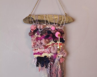 May include: A wall hanging featuring a piece of driftwood with a jute cord for suspension. The textile art is composed of various yarns in shades of pink, magenta, and dark blue, incorporating textured wool, pom-poms, beads, and fringe.