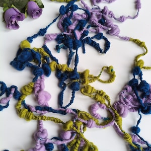 May include: A close-up of colourful, textured yarn in shades of blue, purple, and green. The yarn is arranged in a tangled, organic pattern, with some sections knotted or bunched. Artificial purple roses are in the upper left corner.