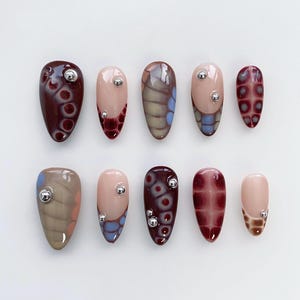 May include: A set of ten artificial nails in various shapes and designs. The nails feature a mix of colors, including burgundy, beige, and brown, with decorative patterns and silver embellishments. The designs range from abstract to geometric.