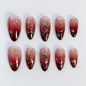 Puede incluir: Un conjunto de diez uñas postizas en forma de almendra. Cada uña presenta un diseño degradado de nude a rojo intenso. Los detalles dorados incluyen soles, lunas, serpientes y estrellas, con pedrería incrustada.