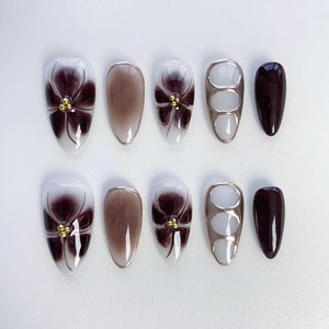 Könnte beinhalten: Satz mit zehn falschen Fingernägeln mit verschiedenen Designs. Einige Nägel haben eine braune Basis mit einem weißen Blumendesign und goldenen Akzenten. Andere Nägel sind einfarbig braun, weiß mit einem goldenen Akzent oder eine klare Basis mit einem goldenen Akzent.
