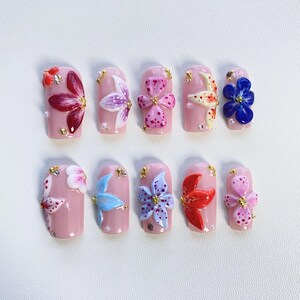 May include: Ten pink nail tips with different flower designs. The flowers are painted in various colours, including red, blue, pink, purple, and white.