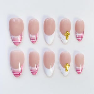 Op de afbeelding: Set van tien kunstnagels in verschillende maten. De nagels hebben een lichtroze basis met een witte punt of een roze ruitpatroon. Eén nagel heeft een gouden kruisbedel. De nagels zijn ovaal van vorm.