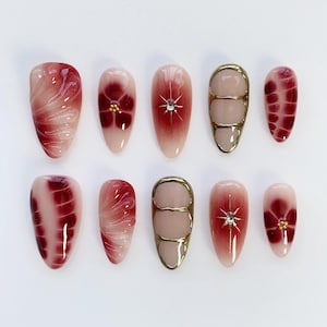 Pode incluir: Um conjunto de dez unhas artificiais com vários designs. As unhas apresentam um gradiente de rosa claro a vermelho escuro, com padrões florais e abstratos. Algumas unhas têm detalhes dourados e strass.