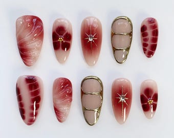 May include: A set of ten artificial nails in various designs. The nails feature a gradient of light pink to deep red, with floral and abstract patterns. Some nails have gold accents and rhinestones.