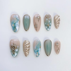 May include: Ten press-on nails with a variety of designs. The nails are a light blue and beige color with gold accents. Some nails feature a gold star, a gold eye, and a gold shell design.