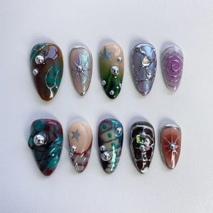 K&ouml;nnte beinhalten: Zehn verschiedene Nagelkunstdesigns mit verschiedenen Farben, Mustern und Verzierungen. Die Designs umfassen einen Wirbel, einen Stern, ein geometrisches Muster und ein Blumenmuster. Jedes Design befindet sich auf einer separaten Nagelform.
