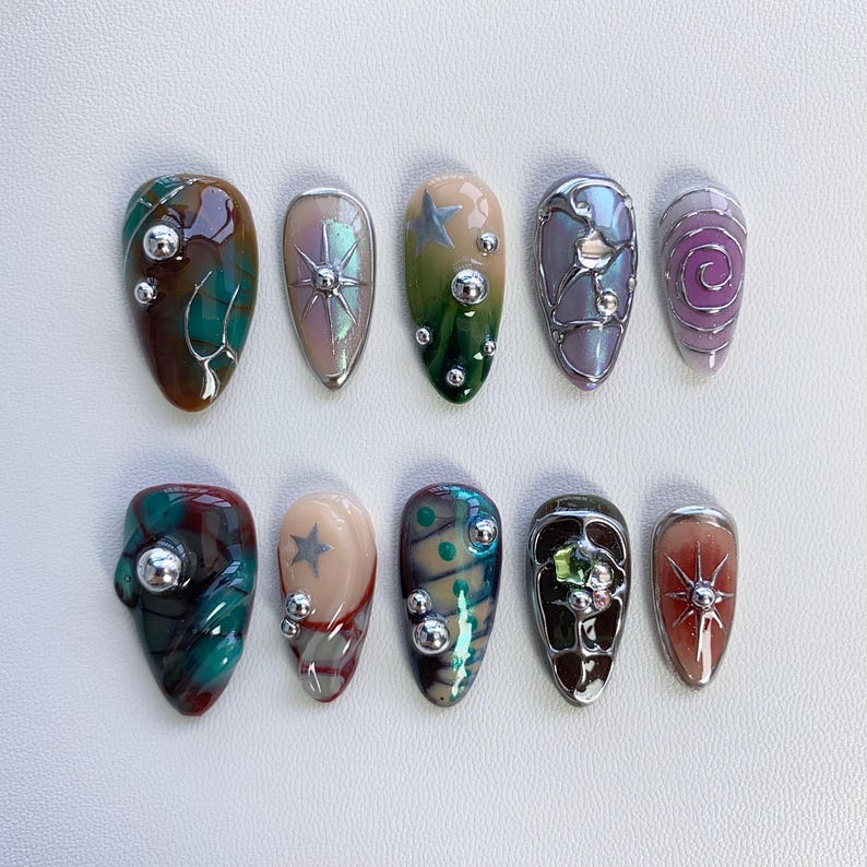 K&ouml;nnte beinhalten: Zehn verschiedene Nagelkunstdesigns mit verschiedenen Farben, Mustern und Verzierungen. Die Designs umfassen einen Wirbel, einen Stern, ein geometrisches Muster und ein Blumenmuster. Jedes Design befindet sich auf einer separaten Nagelform.