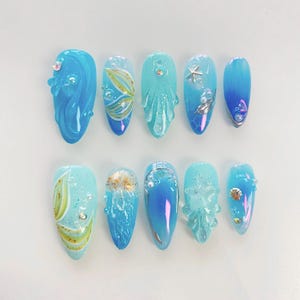 May include: Ten blue and white nail tips with various ocean-themed designs, including a mermaid, seaweed, seashells, and a starfish.