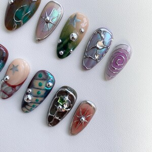 K&ouml;nnte beinhalten: Ein Set mit 12 falschen N&auml;geln mit verschiedenen Designs. Die N&auml;gel sind mit silbernen, goldenen und gr&uuml;nen Akzenten sowie kleinen Perlen und Strasssteinen verziert. Einige der Designs beinhalten einen Stern, einen Wirbel und ein geometrisches Muster.