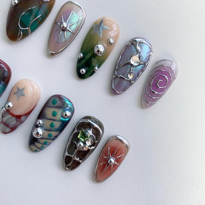 K&ouml;nnte beinhalten: Ein Set mit 12 falschen N&auml;geln mit verschiedenen Designs. Die N&auml;gel sind mit silbernen, goldenen und gr&uuml;nen Akzenten sowie kleinen Perlen und Strasssteinen verziert. Einige der Designs beinhalten einen Stern, einen Wirbel und ein geometrisches Muster.